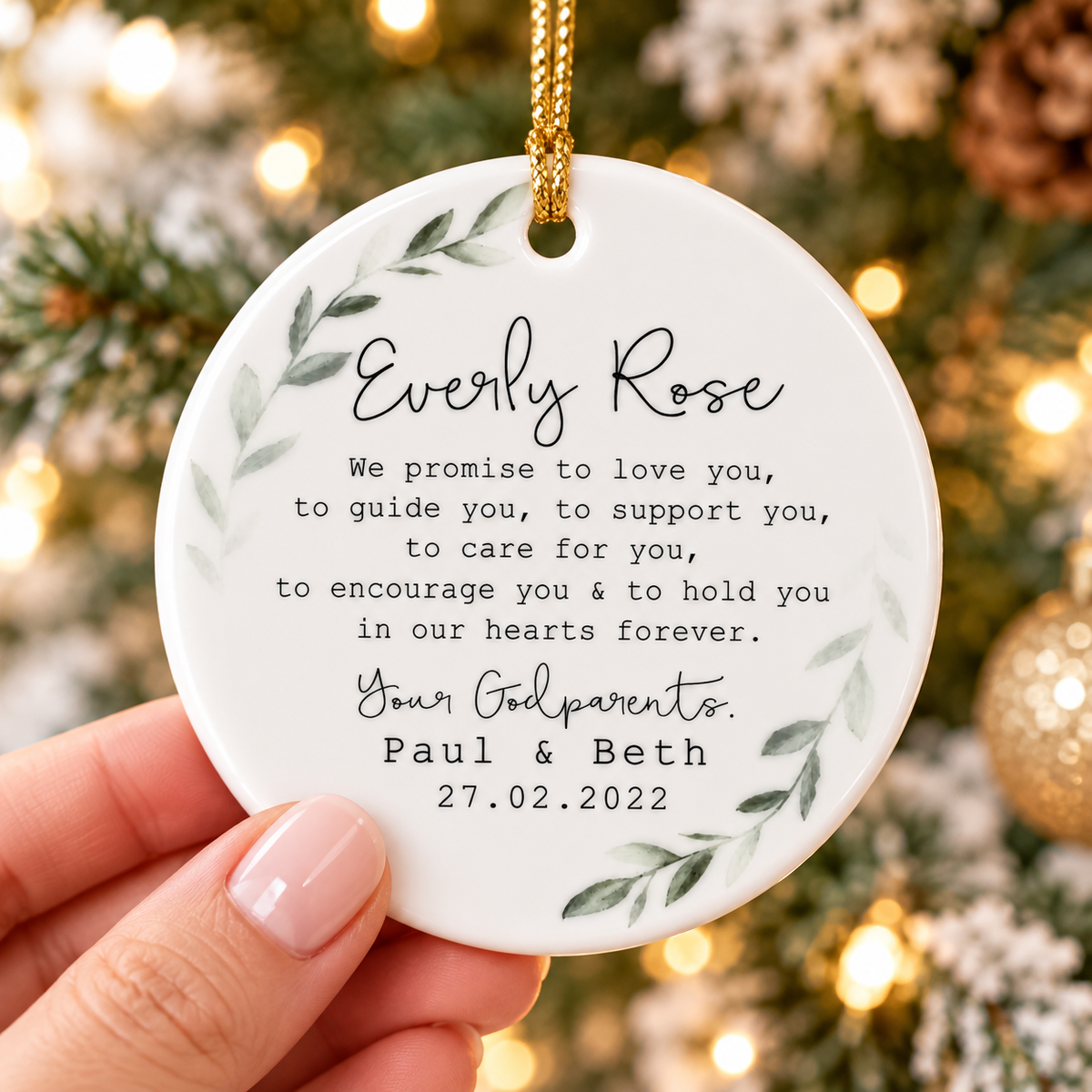 Person holding a round ceramic ornament featuring a green leaf wreath and christening text.