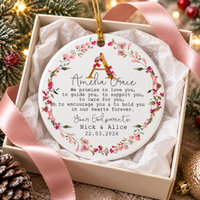 Personalized ceramic ornament inside an open gift box with pink ribbon and pinecone accents.