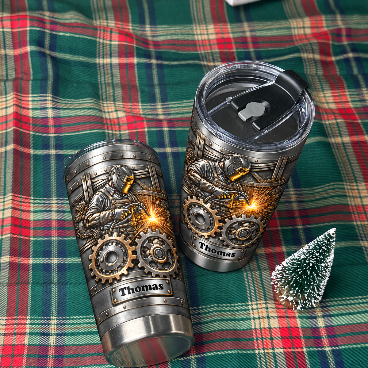 Two steampunk welder tumblers with custom names displayed on a green plaid blanket next to a tiny tree.