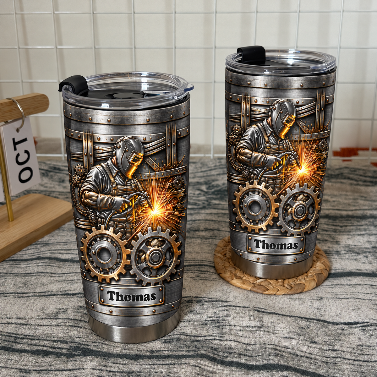 Two vacuum-insulated stainless steel welder tumblers with a steampunk gear design on a rustic textured surface.