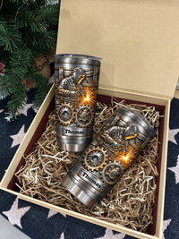 Two custom stainless steel welder tumblers with gears in a gift box with shredded paper, near a tree.