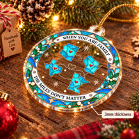 A single clear acrylic holiday reunion gift ornament hanging on a festive pine branch with warm lights.