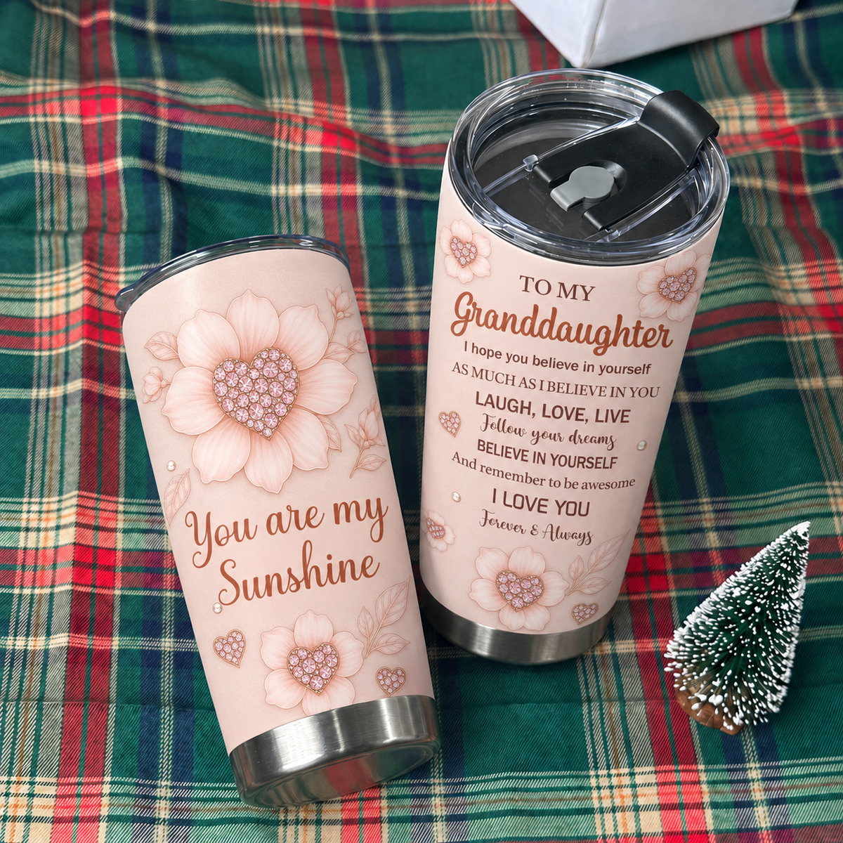 Two inspirational quote tumblers rest on a red and green plaid fabric next to a miniature pine tree.